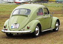 Picture of a yellow automobile in a field, with an airplane in the background. This is a rear three-quarters-view of it.